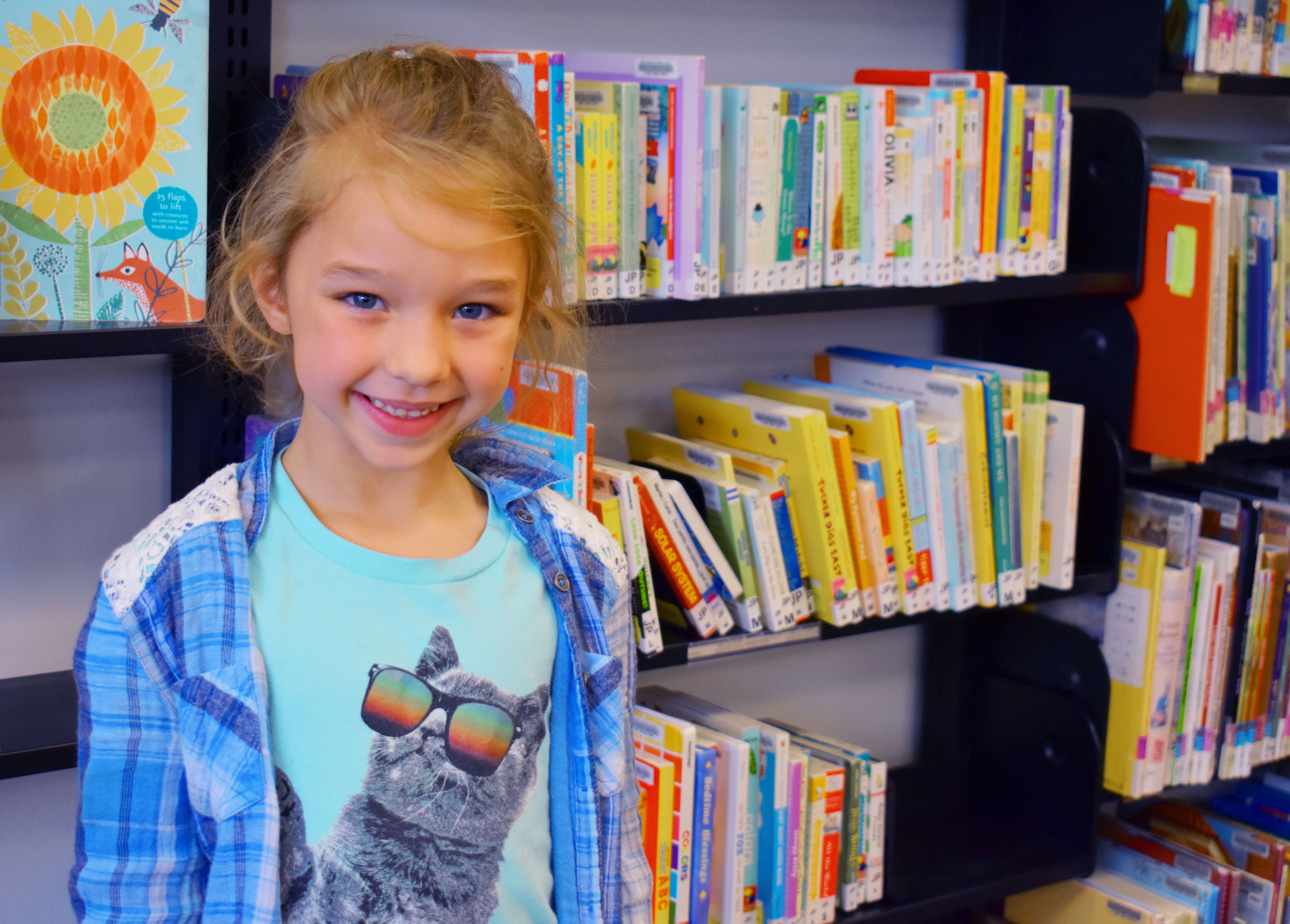 Young girl standing and smiling in front of bookshelves. 