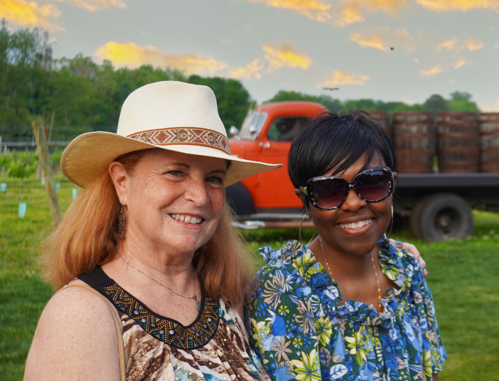 Two women smiling in front of orange flat bed truck in vineyard. 