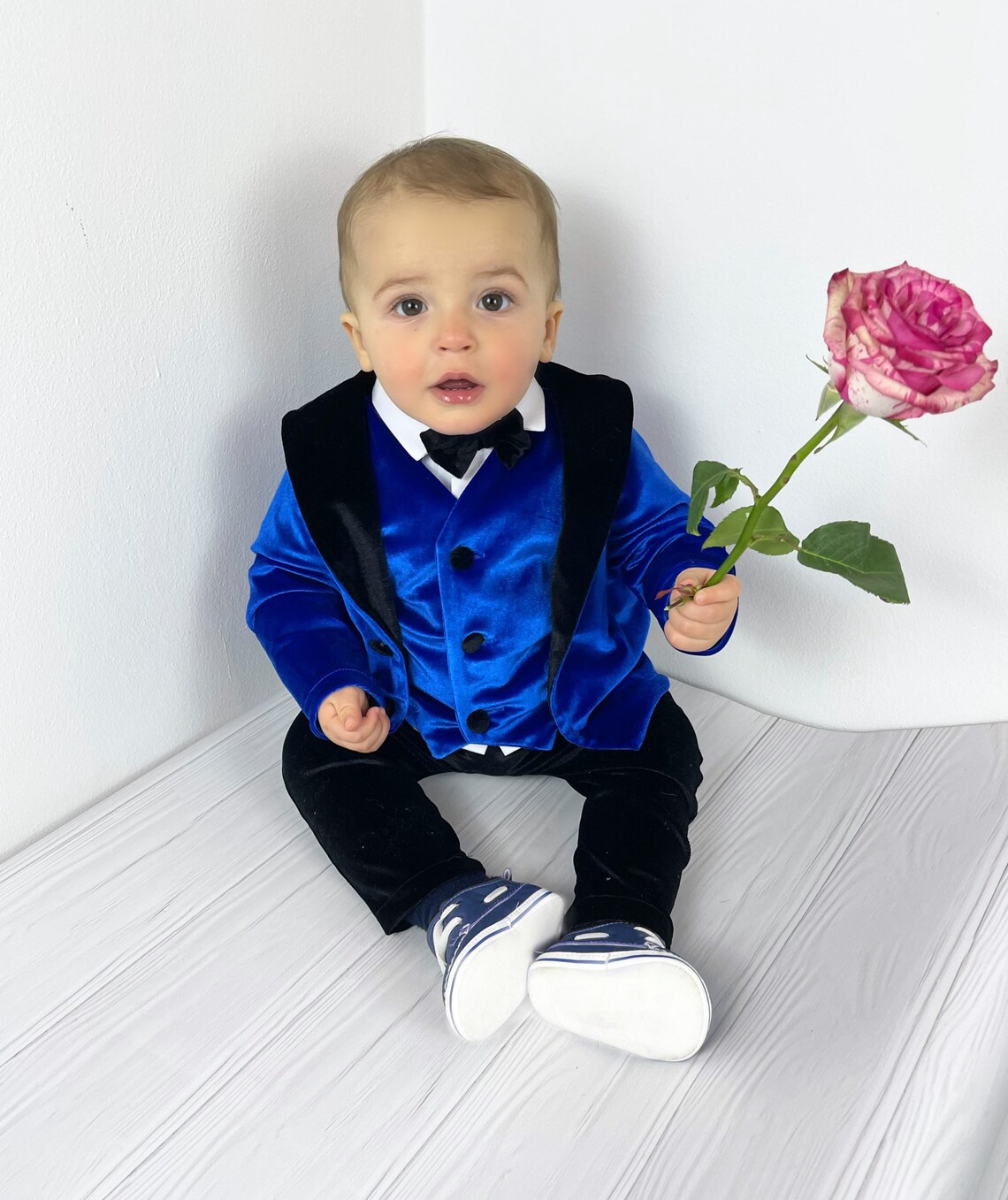 A toddler in a blue tuxedo extends a pink rose toward the screen. He looks very handsome and excited to party at baby prom!