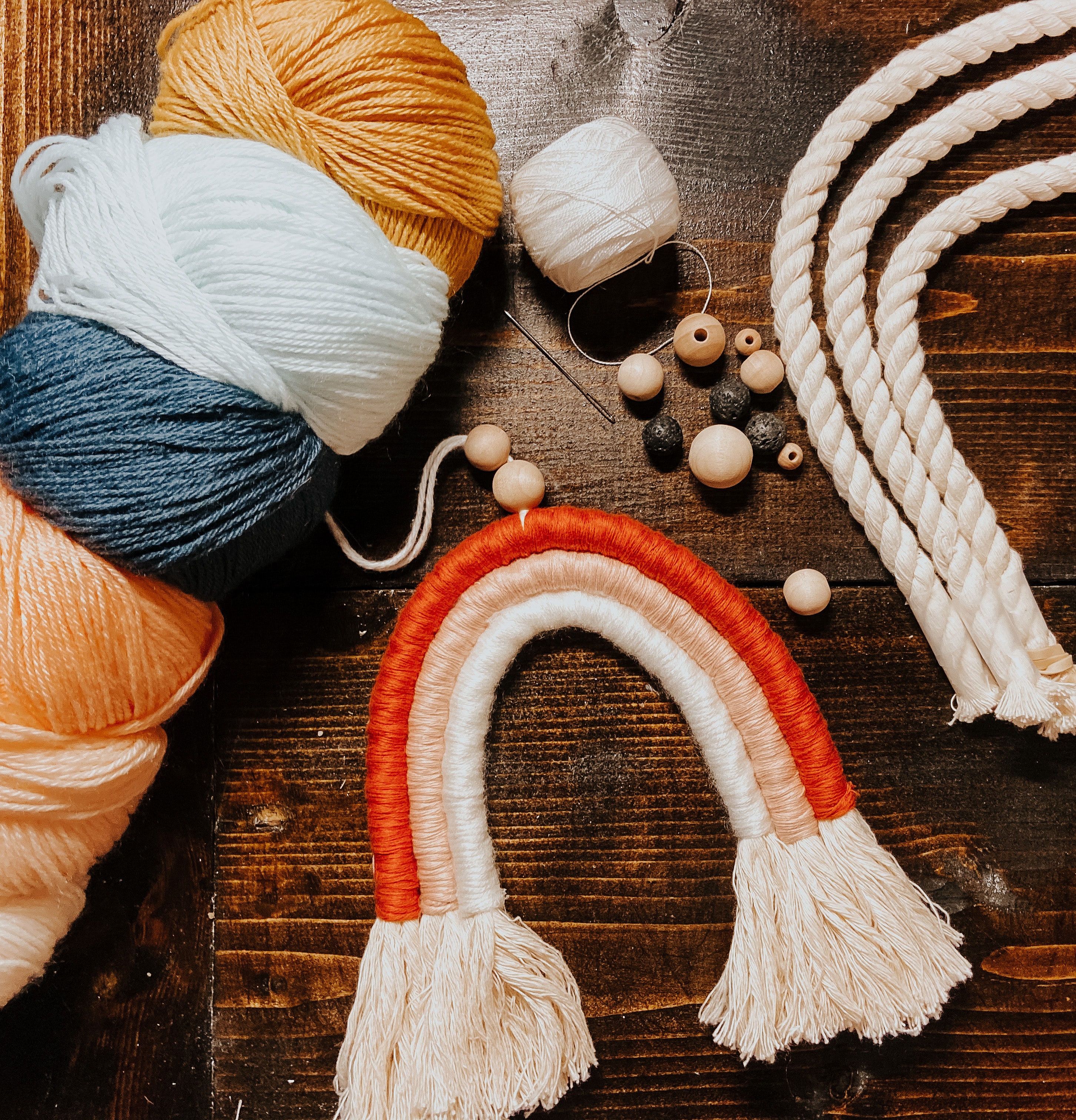 yarn, cord, beads surround a handmade macrame rainbow on a wood table