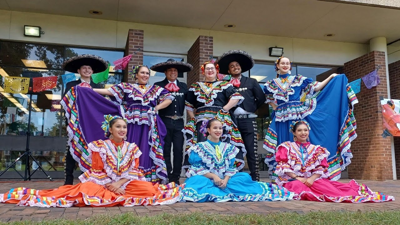 Mexican folk dance group posing for a photo. 3 men standing up, 3 women standing up, and 3 other women sitting down.