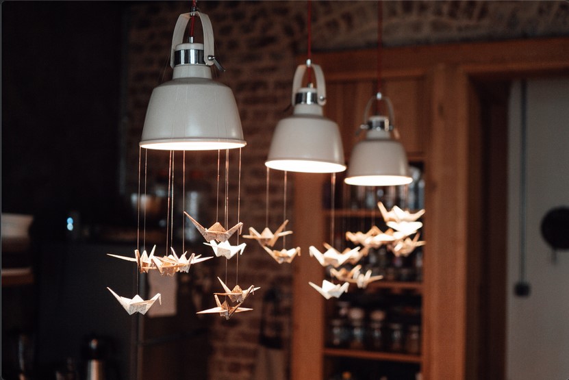 Paper cranes are hung under three hanging lamps and are illuminated with warm light 
