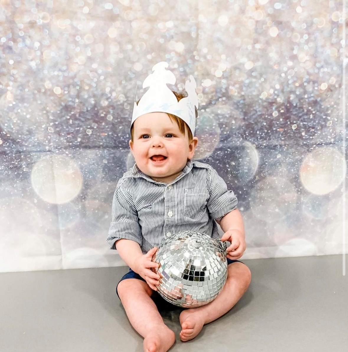 a smiling baby in a crown holds a disco ball in front of a sparkly background