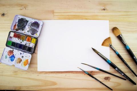 A white canvas, colorful watercolor paint pan, and several brushes are arranged on a wooden background