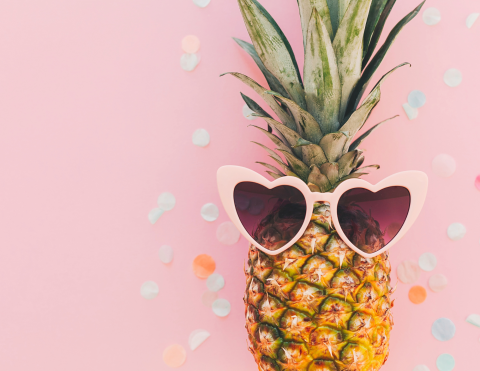 A pineapple wearing heart-shaped glasses and resting on a bed of confetti against a pink background.