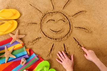 Happy sun finger drawn in the sand by a child. Sun surrounded by a green pair of flip flops, yellow pair of flip flops, towel, and seashells.