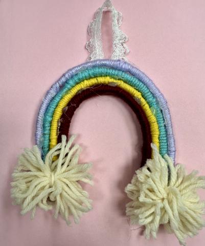 rainbow made with purple, blue, yellow and red yarn. Two white Pom-Poms attached to ends of rainbow