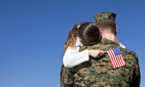 Photo of a young girl holding an American flag while hugging her father. The father is wearing a military uniform.