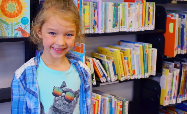 Young girl standing and smiling in front of bookshelves. 